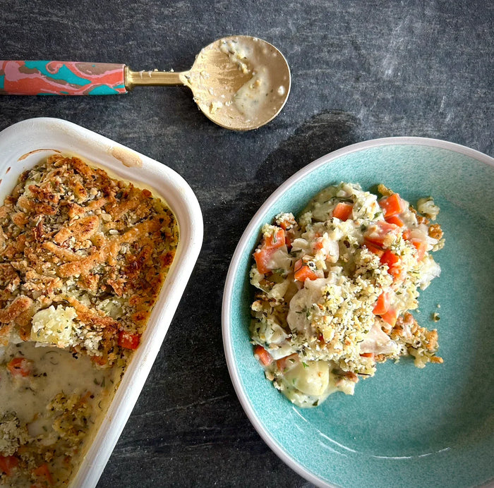 A serving of fish pie with vegetables on a blue plate, next to the remains in a white baking dish, with a spoon on the side.
