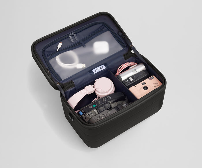 Train case packed with tech gear including multiple cameras, headphones, charging cords, and earpods
